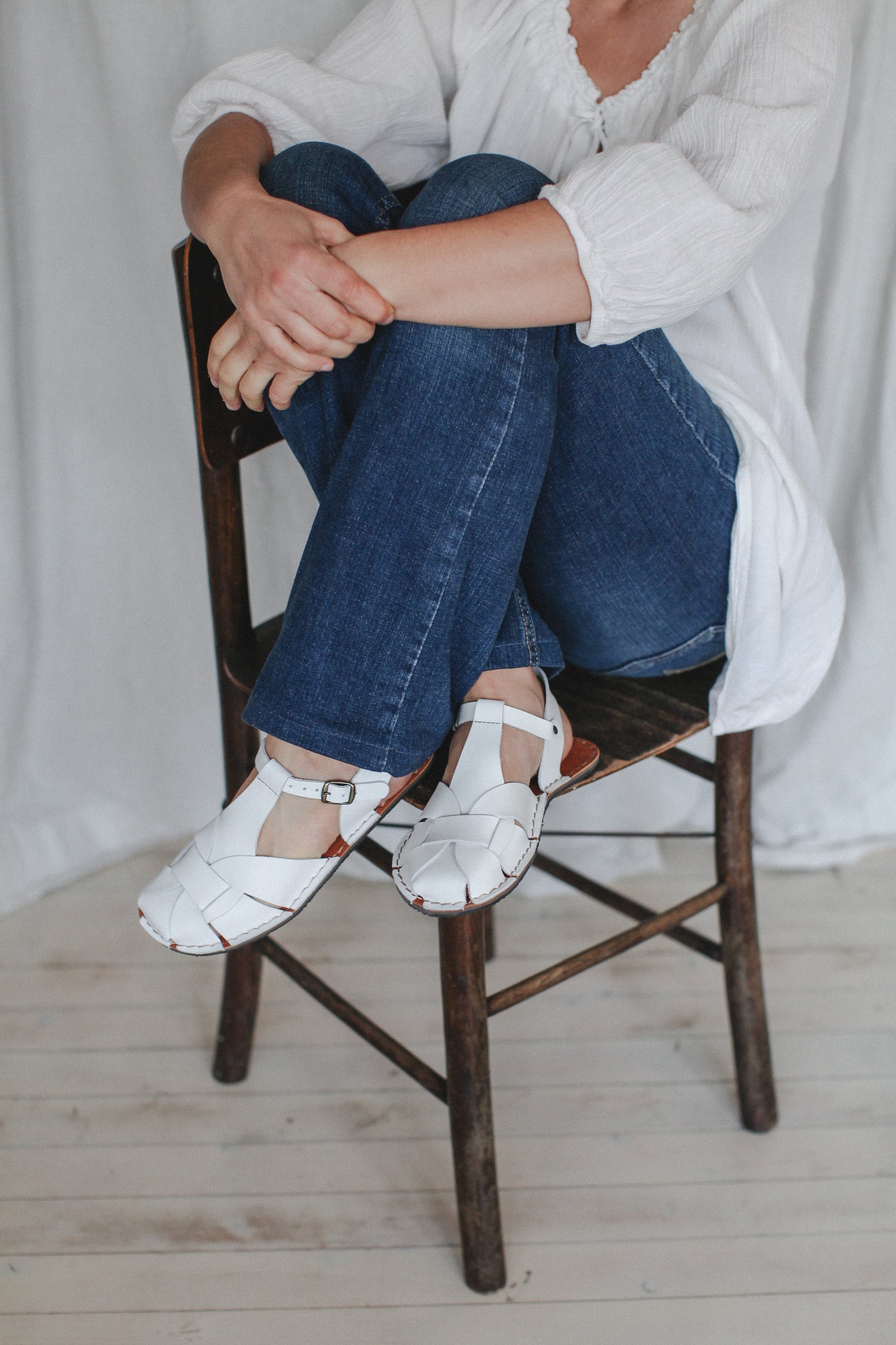 Person sitting on a wooden chair wearing white barefoot sandals, blue jeans, and a white shirt.