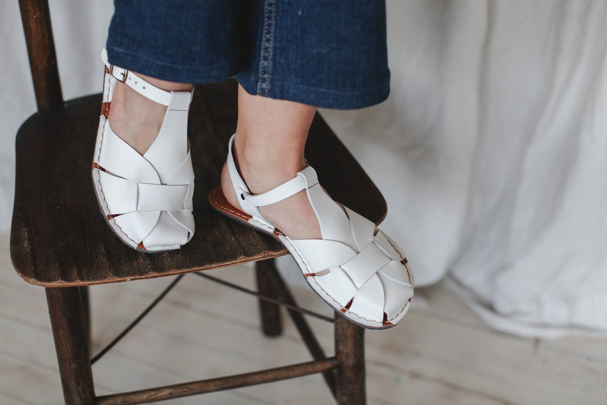 White sandals worn with blue jeans on a wooden stool.